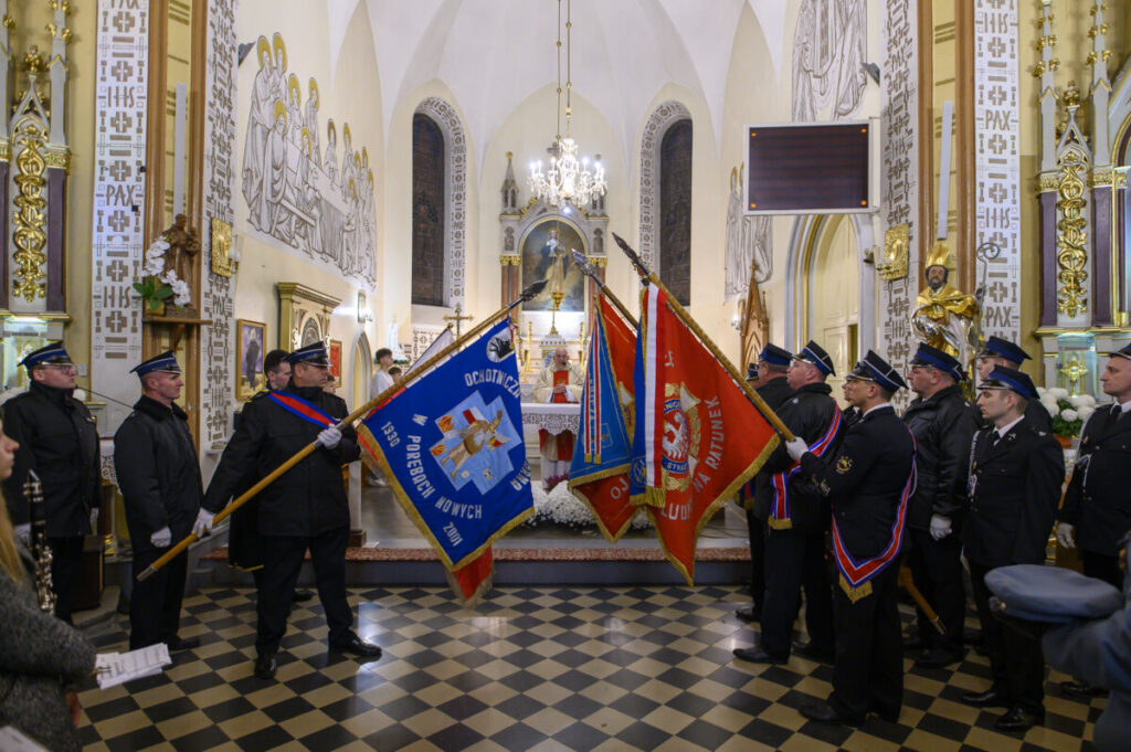 Strażacy w mundurach trzymają sztandary w kościele. Wnętrze jest ozdobne, z dominującymi złotymi i białymi kolorami. Tło to ołtarz i witraże.