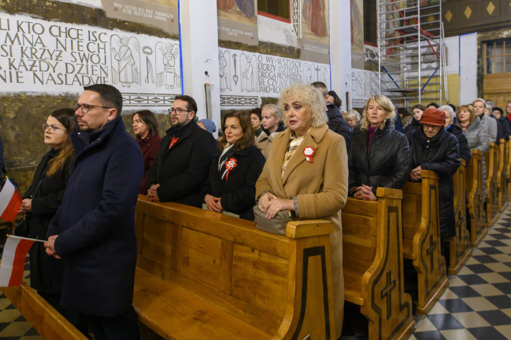 Zdjęcie przedstawia grupę ludzi w kościele podczas ceremonii. Wszyscy stoją w drewnianych ławkach, ubrani na ciemno. W tle widać malowidła ścienne i część ustawionego rusztowania.