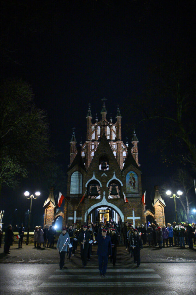 Ceglana kaplica nocą, oświetlona latarniami, otoczona tłumem ludzi. Na fasadzie białe detale i obrazy świętych. Tło ciemne z drzewami.