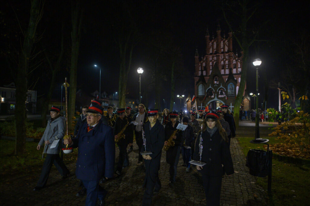 Zespół muzyczny w mundurach maszeruje nocą. Tło to oświetlony kościół z czerwonej cegły. Na zielonym trawniku widać drzewa i latarnie.