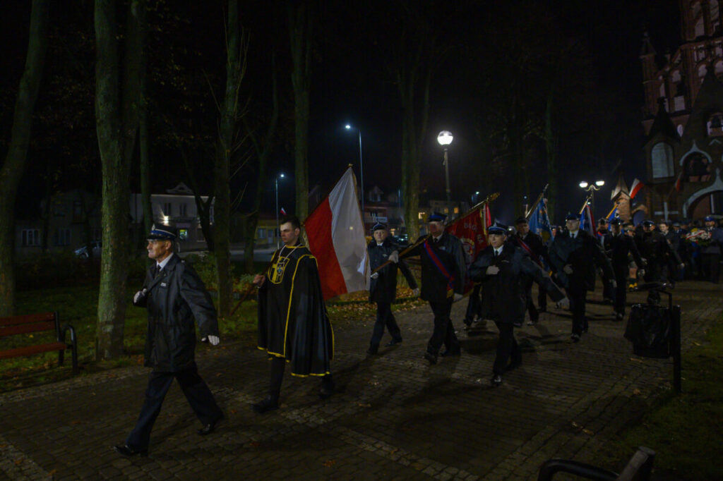 Procesja ludzi w mundurach maszeruje wieczorem z polską flagą. Oświetlone latarnie tworzą ciepły nastrój. W tle drzewa i budynek.