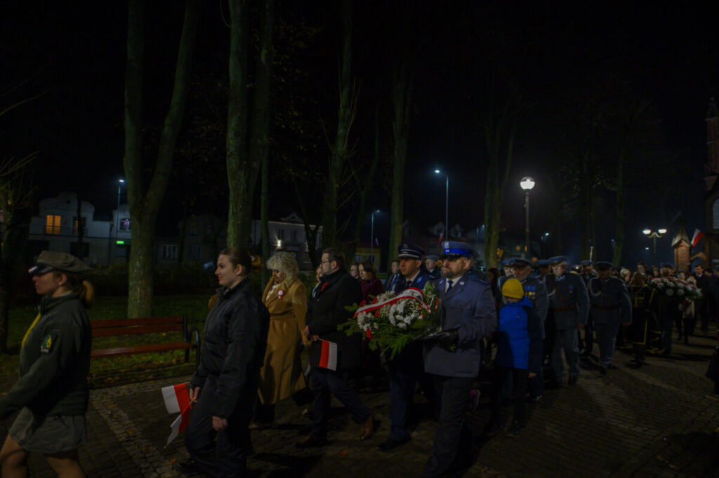 Grupa ludzi, w tym osoby w mundurach, niesie wieniec w parku nocą. Światła latarni oświetlają ich kroki. Tło jest ciemne z widocznym drzewami.