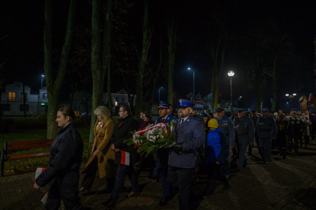 Grupa ludzi maszeruje nocą w parku, niosąc wieńce. Ubrani w ciemne płaszcze i mundury, w tle drzewa i latarnie. Widoczne polskie flagi i symbolika patriotyczna.