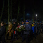 Grupa ludzi maszeruje nocą w parku, niosąc wieńce. Ubrani w ciemne płaszcze i mundury, w tle drzewa i latarnie. Widoczne polskie flagi i symbolika patriotyczna.