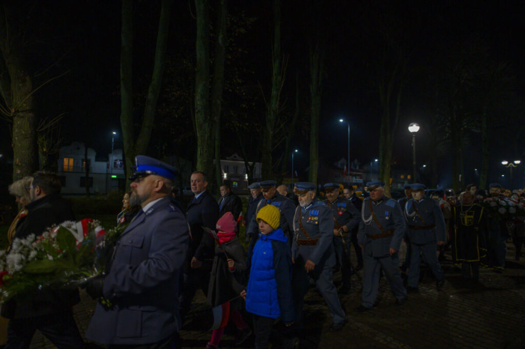 Ludzie w mundurach i cywile uczestniczą w nocnej paradzie. Tło to oświetlona ulica z drzewami i budynkami. Dzieci w kolorowych ubraniach idą z dorosłymi.