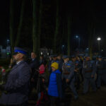 Ludzie w mundurach i cywile uczestniczą w nocnej paradzie. Tło to oświetlona ulica z drzewami i budynkami. Dzieci w kolorowych ubraniach idą z dorosłymi.