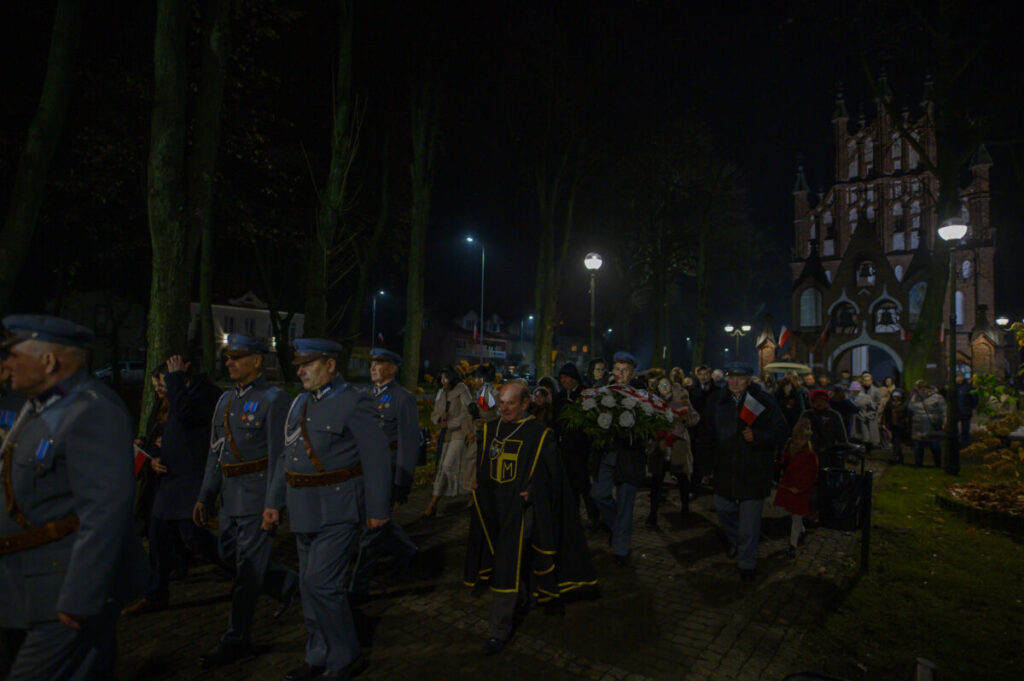 Wieczorna procesja przy kościele z czerwonej cegły. Ludzie w mundurach i stroje cywilnej dekorują kwiatami. W tle drzewa i latarnie.