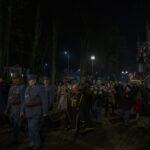 Wieczorna procesja przy kościele z czerwonej cegły. Ludzie w mundurach i stroje cywilnej dekorują kwiatami. W tle drzewa i latarnie.