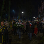 Grupa ludzi maszeruje nocą, trzymając kwiaty i flagi. W tle oświetlony, ceglasty kościół. Otoczenie parku, ciemne kolory.