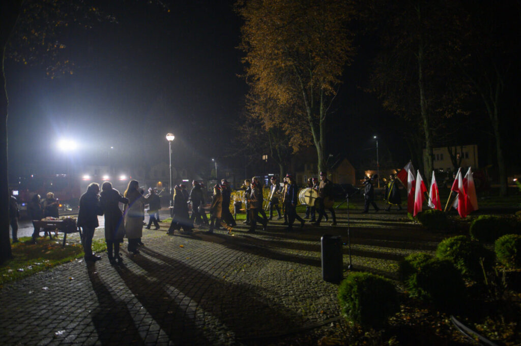 Na zdjęciu nocą marsz ulicą, w tle polskie flagi. Tłum ludzi idzie oświetlony latarniami. Otoczenie parku, drzewa jesienią.