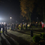 Na zdjęciu nocą marsz ulicą, w tle polskie flagi. Tłum ludzi idzie oświetlony latarniami. Otoczenie parku, drzewa jesienią.