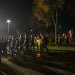 Nocny marsz z flagą Polski i orkiestrą dętą idącą ulicą. W tle drzewa i latarnia. Uczestnicy w ciemnych ubraniach.
