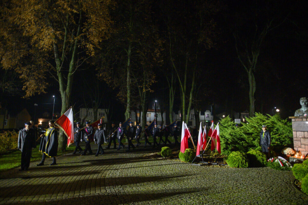 Wieczorna uroczystość z flagami Polski. Ludzie maszerują, otoczeni drzewami w parku. Pomnik i znicze widoczne na tle.