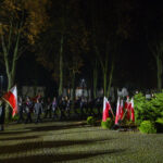Wieczorna uroczystość z flagami Polski. Ludzie maszerują, otoczeni drzewami w parku. Pomnik i znicze widoczne na tle.
