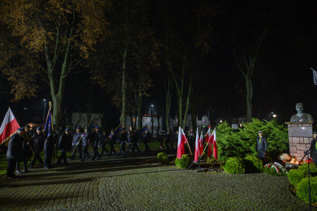 Grupa ludzi maszeruje z flagami w parku nocą. Tło z drzewami i budynkami. Pomnik otoczony biało-czerwonymi flagami.
