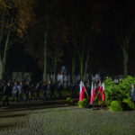 Grupa ludzi maszeruje z flagami w parku nocą. Tło z drzewami i budynkami. Pomnik otoczony biało-czerwonymi flagami.