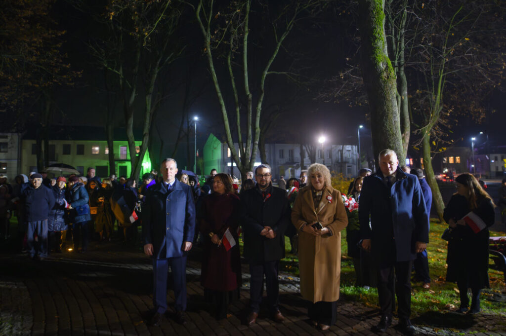 Grupa ludzi stoi na placu nocą. W tle widoczne drzewa i budynki, oświetlone reflektorami. Uczestnicy trzymają flagi Polski.