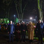 Grupa ludzi stoi na placu nocą. W tle widoczne drzewa i budynki, oświetlone reflektorami. Uczestnicy trzymają flagi Polski.