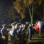 Grupa ludzi w mundurach salutuje na zewnątrz nocą. Flagi polskie na pierwszym planie, w tle drzewa i budynki. Oświetlenie ciepłe, deszcz.