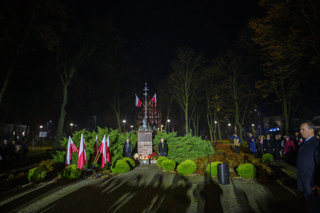 Nocna uroczystość przy pomniku, otoczonym zielenią i flagami Polski. W tle park z drzewami i spacerującymi ludźmi.