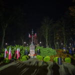 Nocna uroczystość przy pomniku, otoczonym zielenią i flagami Polski. W tle park z drzewami i spacerującymi ludźmi.
