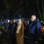 Grupa ludzi stoi na zewnątrz nocą. Mają na sobie płaszcze, w tle drzewa i światła uliczne. Uczestniczą w wydarzeniu.