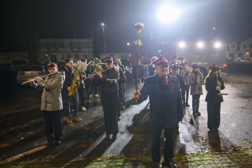 Orkiestra marszowa w mundurach gra instrumenty na oświetlonym placu. Nocne tło z budynkami i samochodami. Dominują ciemne kolory.