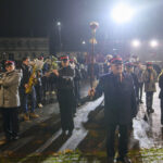 Orkiestra marszowa w mundurach gra instrumenty na oświetlonym placu. Nocne tło z budynkami i samochodami. Dominują ciemne kolory.