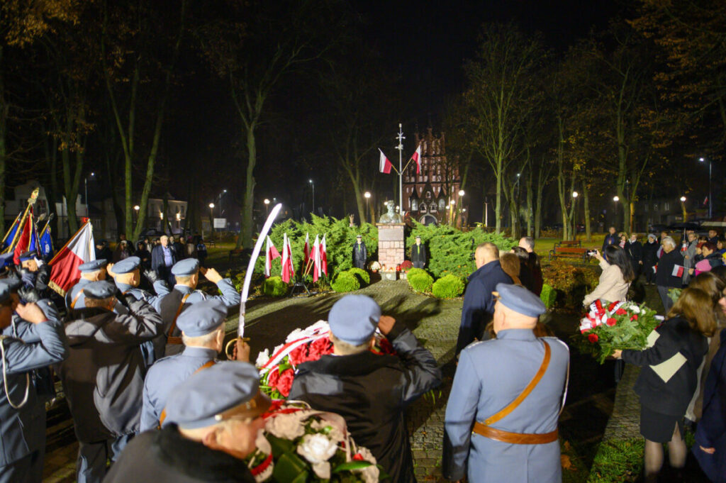 Na zdjęciu nocna uroczystość przy pomniku z flagami. Ludzie w uniformach salutują, trzymając wieńce. W tle drzewa i budynek.