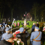 Na zdjęciu nocna uroczystość przy pomniku z flagami. Ludzie w uniformach salutują, trzymając wieńce. W tle drzewa i budynek.