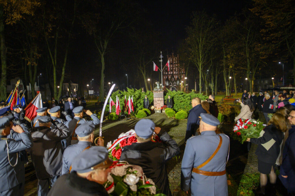 Grupa ludzi oddaje hołd przy pomniku w nocnym parku. Ubrani na granatowo, w tle widoczne są polskie flagi i kościół. Drzewa otaczają plac, tworząc uroczystą atmosferę.