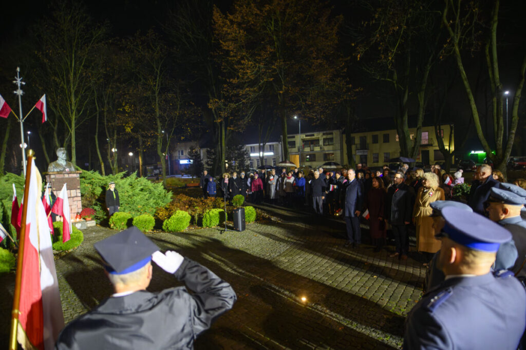 Na zdjęciu nocna uroczystość z tłumem ludzi, żołnierz salutuje. W tle zieleń i wizerunek popiersia. Flagi biało-czerwone.