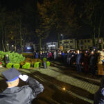 Na zdjęciu nocna uroczystość z tłumem ludzi, żołnierz salutuje. W tle zieleń i wizerunek popiersia. Flagi biało-czerwone.