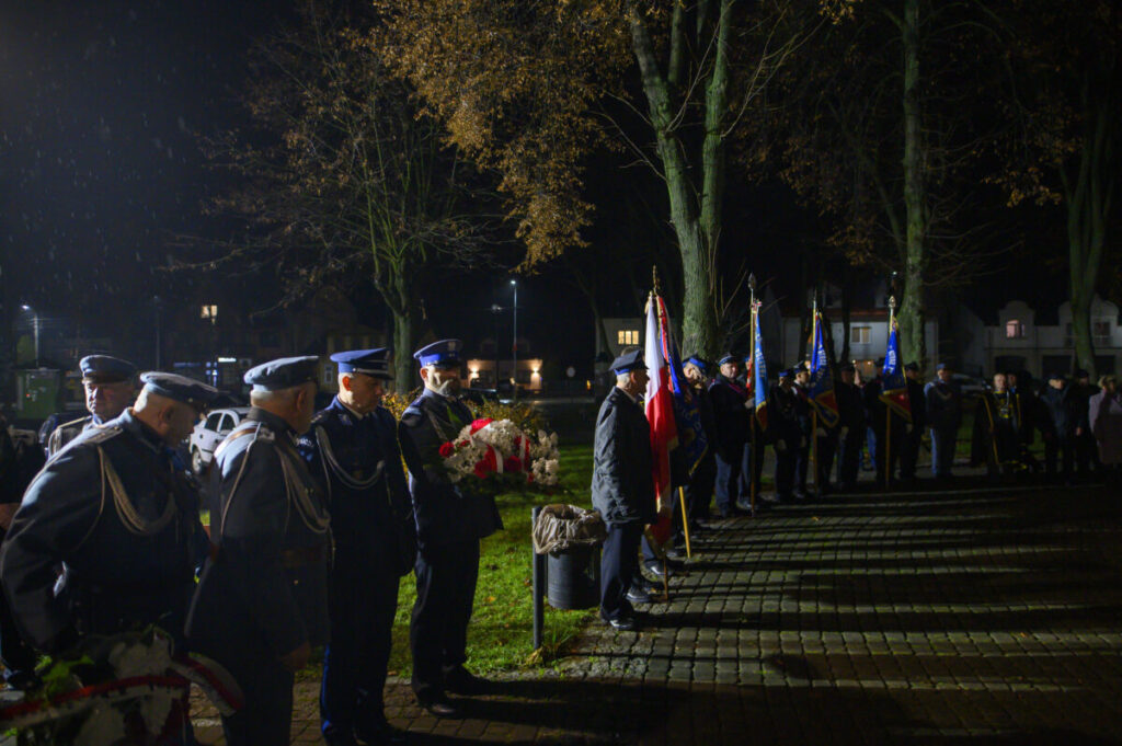 Grupa umundurowanych ludzi stoi w nocnym parku, trzymając flagi i wieńce. Dominuje ciemna kolorystyka z oświetleniem lamp ulicznych. Tło stanowią drzewa i budynki.