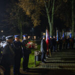 Grupa umundurowanych ludzi stoi w nocnym parku, trzymając flagi i wieńce. Dominuje ciemna kolorystyka z oświetleniem lamp ulicznych. Tło stanowią drzewa i budynki.