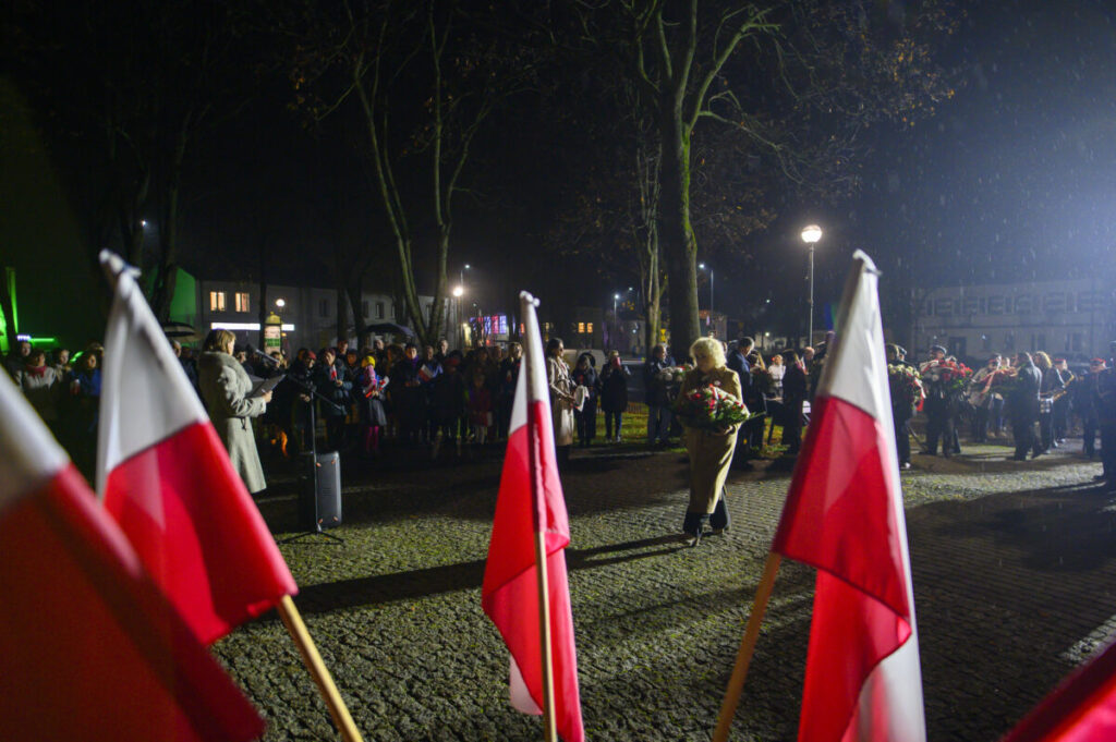 Grupa ludzi na wieczorze, w tle ciemne niebo i drzewa. Flagi polskie dominują na pierwszym planie. Postać z kwiatami idzie przed tłum.
