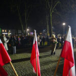 Grupa ludzi na wieczorze, w tle ciemne niebo i drzewa. Flagi polskie dominują na pierwszym planie. Postać z kwiatami idzie przed tłum.