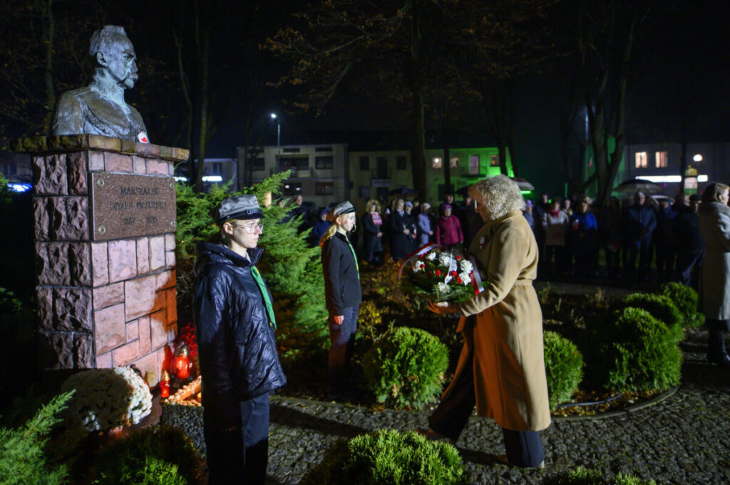 Pomnik Józefa Piłsudskiego nocą, otoczony zielenią. Kobieta składa wieniec, przygląda się jej tłum w tle. Jasne oświetlenie.