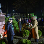 Pomnik Józefa Piłsudskiego nocą, otoczony zielenią. Kobieta składa wieniec, przygląda się jej tłum w tle. Jasne oświetlenie.