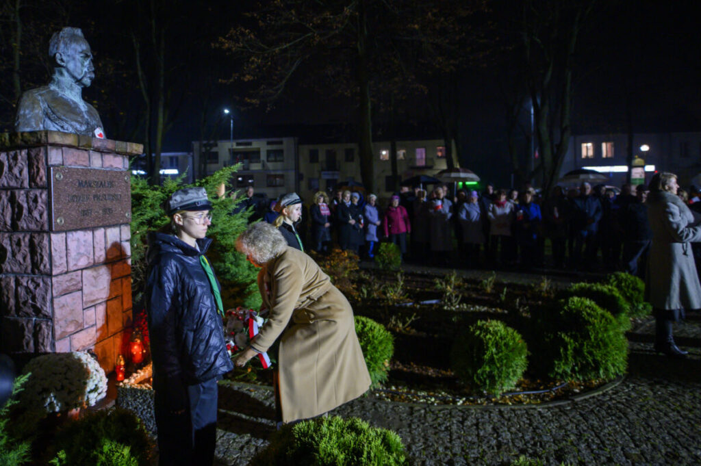 Pomnik w formie popiersia na tle nocnym, ludzie składają wieniec. Wokół świeci się czerwone światło zniczy, a w oddali stoi tłum.