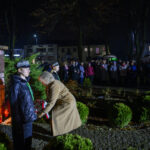 Pomnik w formie popiersia na tle nocnym, ludzie składają wieniec. Wokół świeci się czerwone światło zniczy, a w oddali stoi tłum.