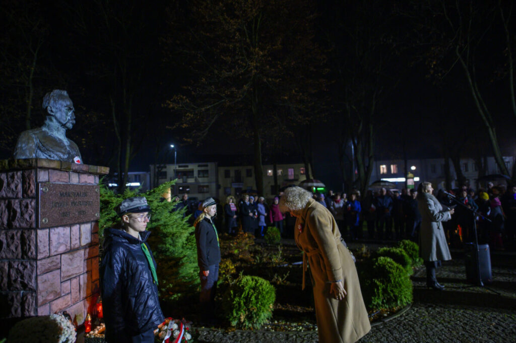 Bokiem pomnik na tle nocnego parku, wokół grupa ludzi, czerwono-zielone oświetlenie. Osoby w jasnych płaszczach z przodu.