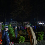 Bokiem pomnik na tle nocnego parku, wokół grupa ludzi, czerwono-zielone oświetlenie. Osoby w jasnych płaszczach z przodu.