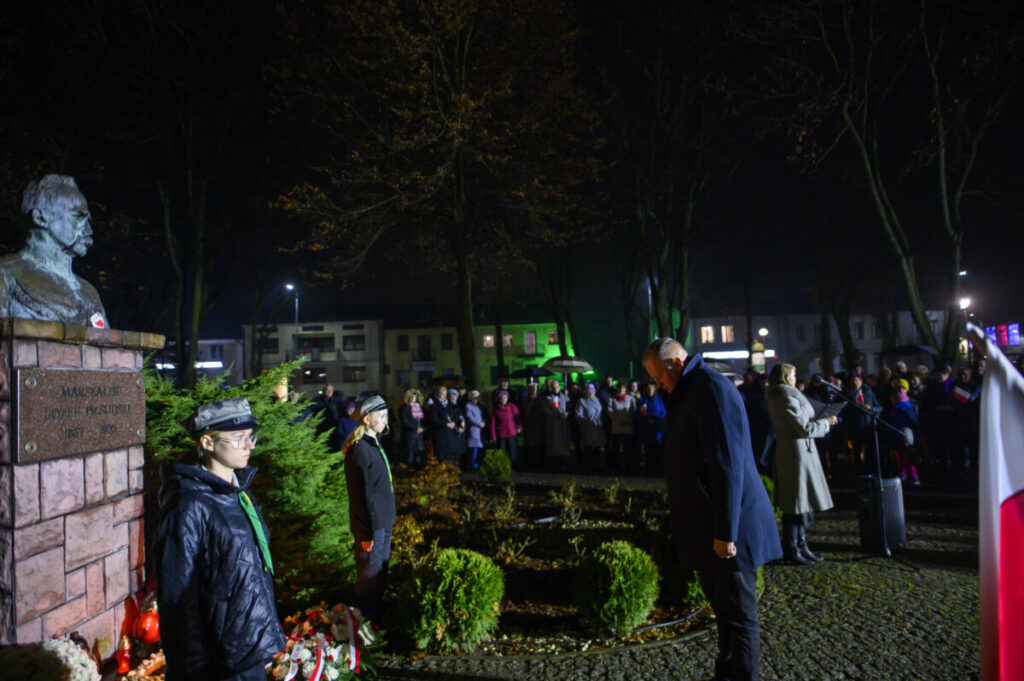 Pomnik Piłsudskiego nocą otoczony przez ludzi. Oświetlenie zielone i czerwone, z białą flagą po prawej. Drzewa i budynki w tle.
