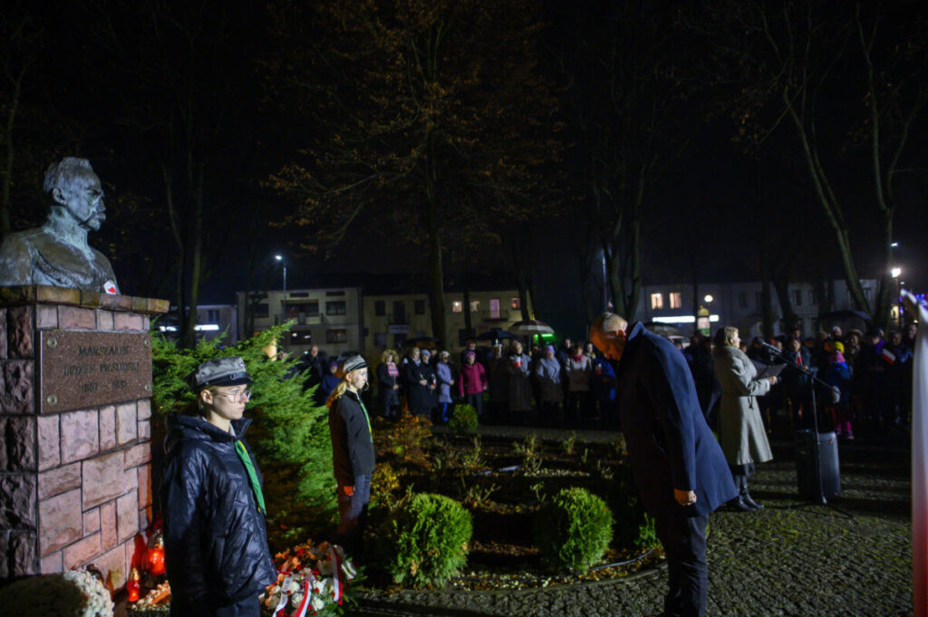 Pomnik marszałka Piłsudskiego na tle parku nocą. Ludzie zgromadzeni w ciemnych ubraniach, wokół kwiaty i znicze. Tło ciemne z ciepłym oświetleniem.