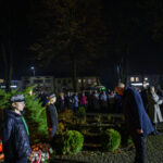 Pomnik marszałka Piłsudskiego na tle parku nocą. Ludzie zgromadzeni w ciemnych ubraniach, wokół kwiaty i znicze. Tło ciemne z ciepłym oświetleniem.