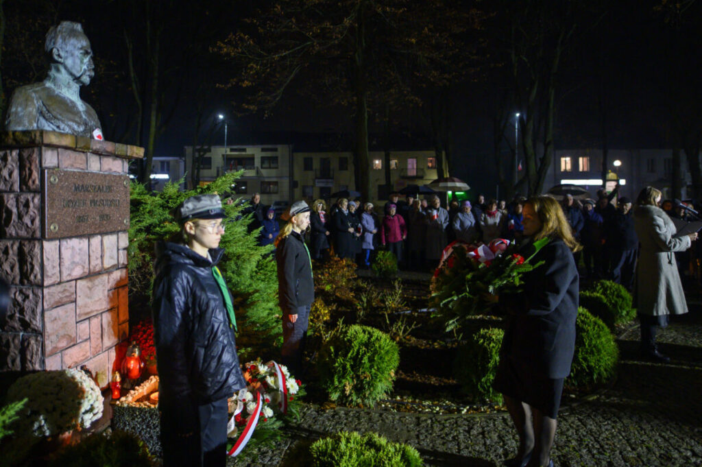Pomnik marszałka Piłsudskiego w parku nocą. Ludzie składają kwiaty, otoczenie oświetlone sztucznym światłem. Tło to ciemne drzewa i budynki.