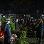 Pomnik marszałka Piłsudskiego w parku nocą. Ludzie składają kwiaty, otoczenie oświetlone sztucznym światłem. Tło to ciemne drzewa i budynki.