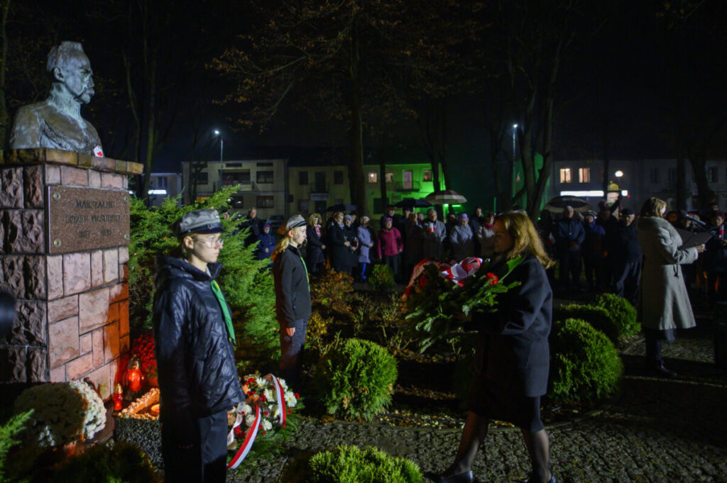 Pomnik z popiersiem marszałka otoczony zielenią, z ludźmi składającymi wieńce przy nim. Jasne światło oświetla scenę nocą, tworząc kontrast z ciemnym tłem. Wydarzenie gromadzi tłum osób w zimowych ubraniach