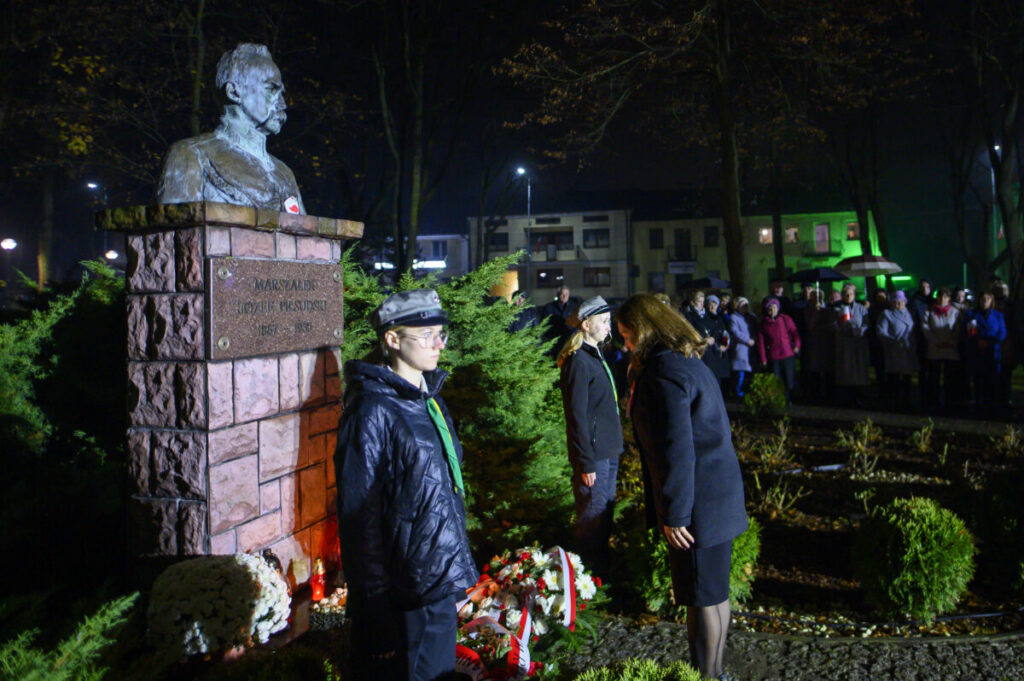 Pomnik Piłsudskiego nocą, otoczony zielenią. Grupa ludzi w tle, światła i wieńce. Uroczystość upamiętniająca.